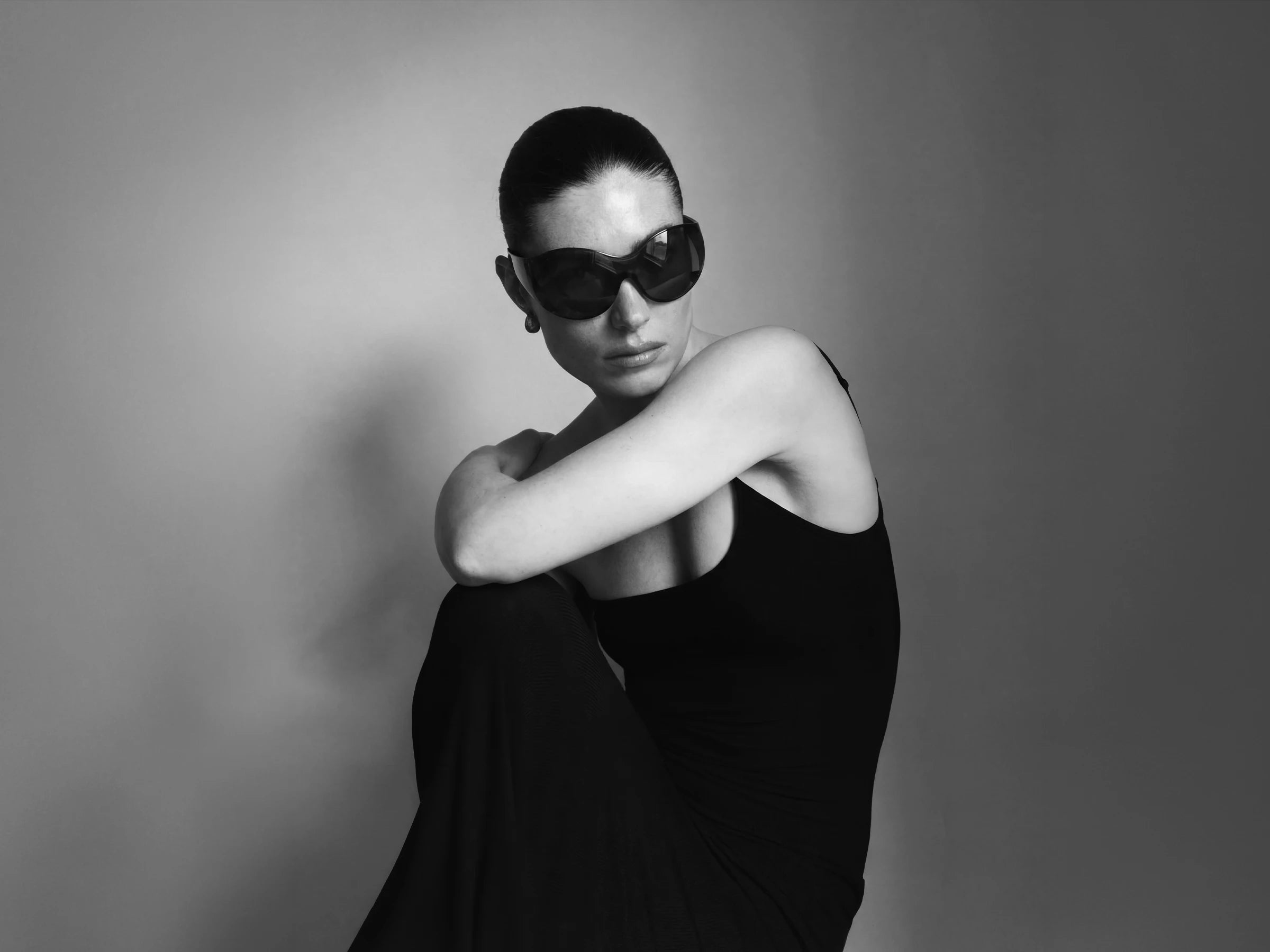 Black and white portrait of a person wearing theflyshades sunglasses and a black dress against a plain background