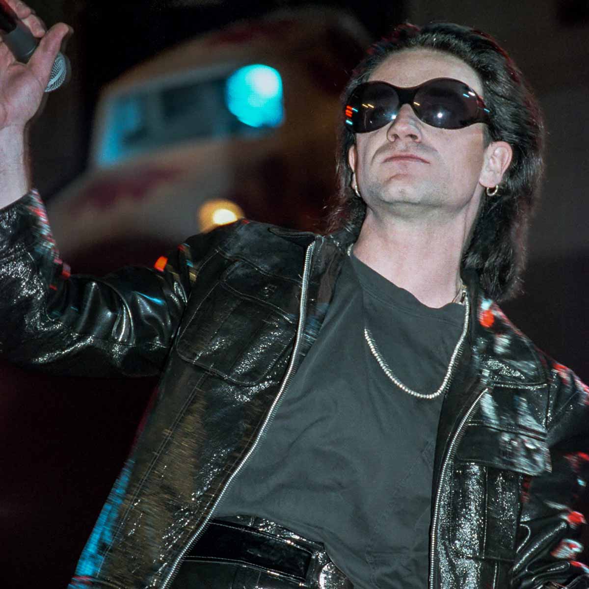 Bono wearing a black leather jacket and Theflyshades sunglasses on stage with a blurred background